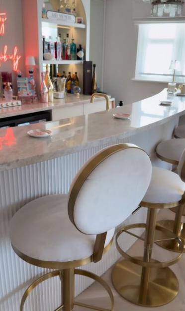 A home bar with a marble countertop, white and gold bar stools, a neon sign, and shelves stocked with various liquor bottles.