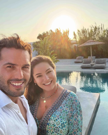 A man and a woman smiling for a selfie by a pool at sunset.