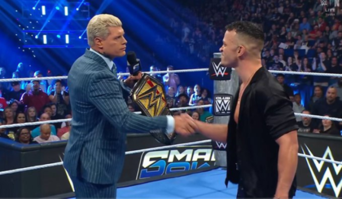 Cody Rhodes in a blue suit shaking hands with another man in a black shirt in a wrestling ring.
