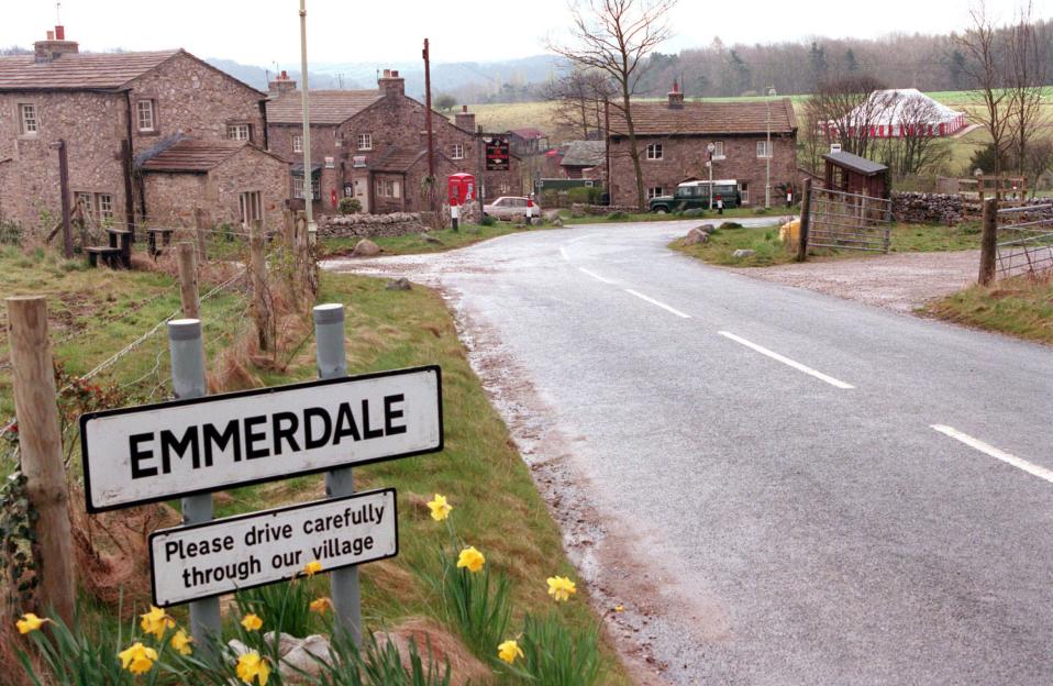 A road sign for Emmerdale village with stone houses and a red phone booth in the background.