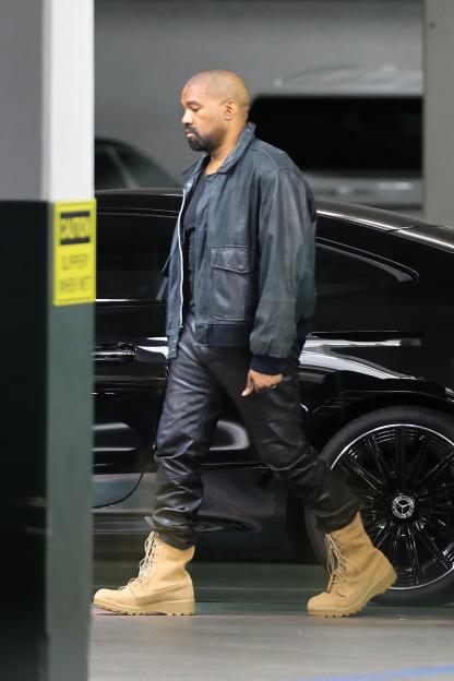 Kanye West in a black leather jacket, black leather pants, and tan boots, walking by a black car.