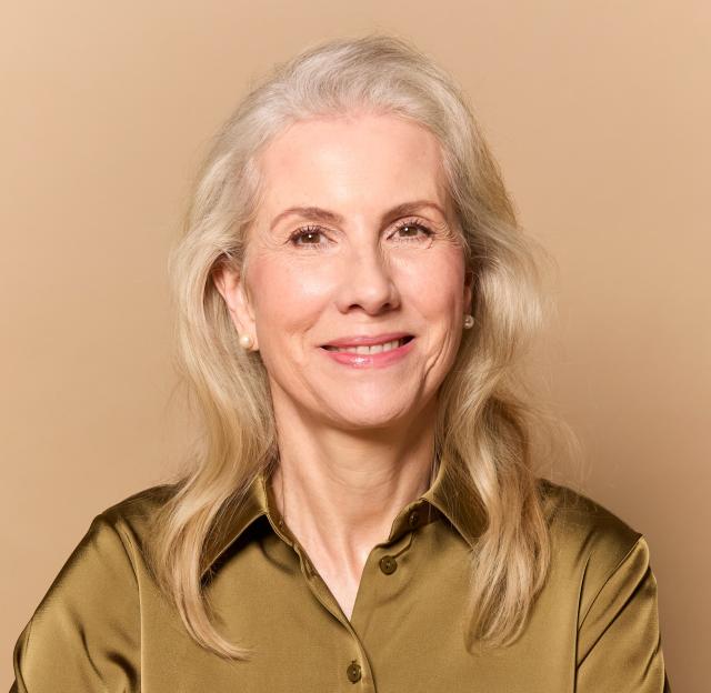Katrina Verdon-Roe, 61, sits at a table and smiles at the camera.