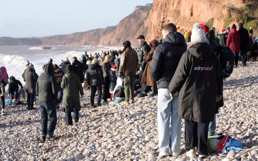 A large group of people gather on a rocky beach, watching waves crash on the shore, with cliffs in the background.