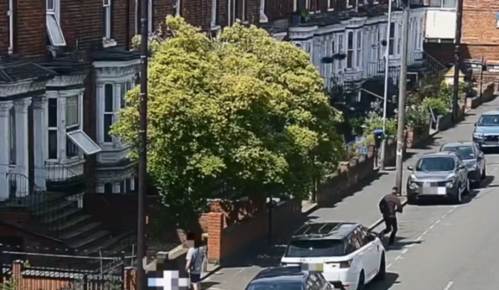 A man in dark clothing running along a street with parked cars, and a tree in front of a building.