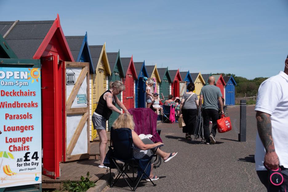 People on a beach by colorful beach huts with a sign advertising deckchair rentals.