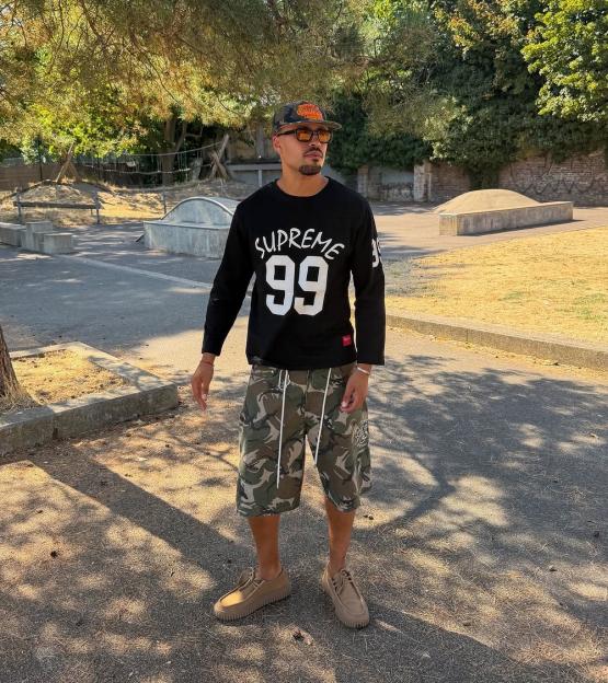 Man in a Supreme 99 shirt, camouflage shorts, and tan shoes standing outdoors.