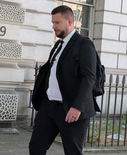 PC Daniel Tomkins in a black suit with a backpack walking outside a building.