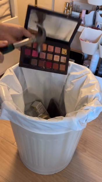 Person destroying a Huda Beauty eyeshadow palette with a hammer over a trash can.