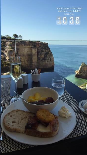 A breakfast plate with toast, bacon, hash brown, a bowl of fruit with chia seeds, a glass of champagne, and a glass of water on a table overlooking a cliff and the ocean.