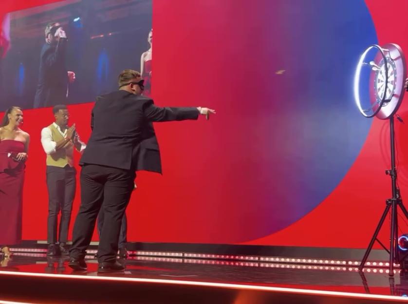 Luke Littler blindfolded, throwing a dart at a dartboard on stage.