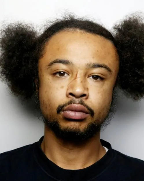 A headshot of Shaylon McCalla with dark skin, dark curly hair, a beard, and a visible scar across his nose.