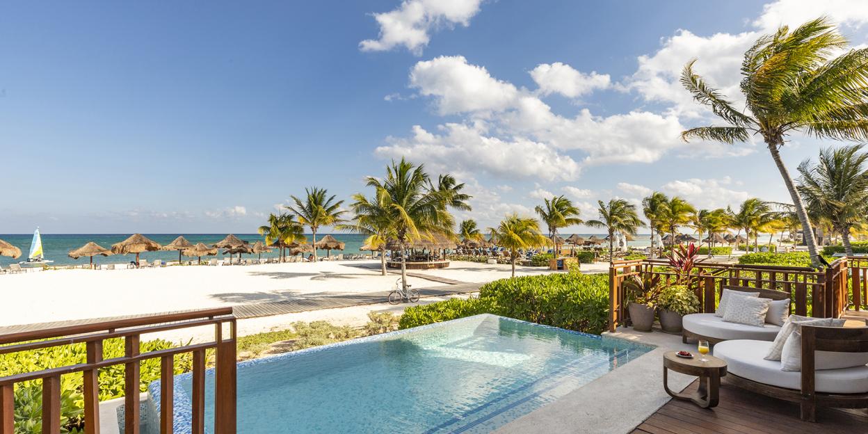 Luxury hotel with a patio and pool overlooking a white sand beach with palm trees and cabanas.