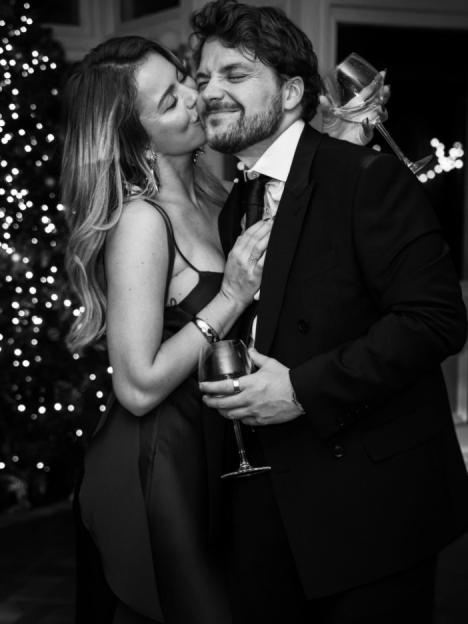 A woman in a slip dress kissing a man in a suit on the cheek, both holding wine glasses, with a blurred Christmas tree in the background.