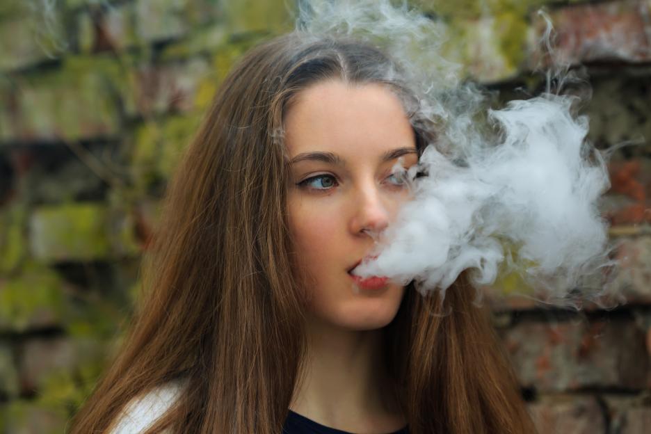 A young woman with long brown hair exhales a large cloud of vape smoke, looking towards the right, with a blurred brick wall in the background.