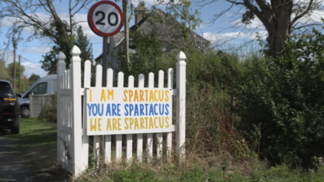 A sign on a white picket fence reads "I am Spartacus, you are Spartacus, we are Spartacus."