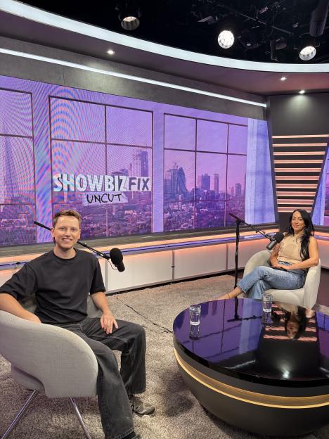 Two hosts, a man and a woman, seated in a studio with a city skyline backdrop and a "SHOWBIZFIX UNCUT" logo.