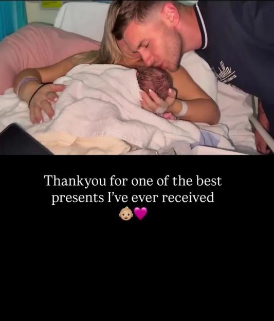 A man kisses a newborn baby held by a woman in a hospital bed, with text wishing his wife a happy anniversary.