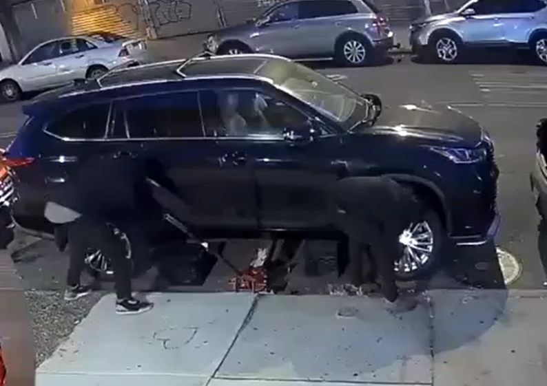 Two individuals work to remove parts from a black SUV, which is lifted by a jack.