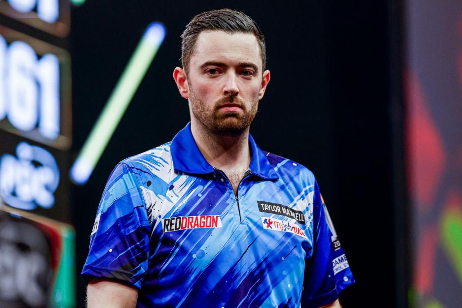 A male darts player in a blue shirt with various sponsors.