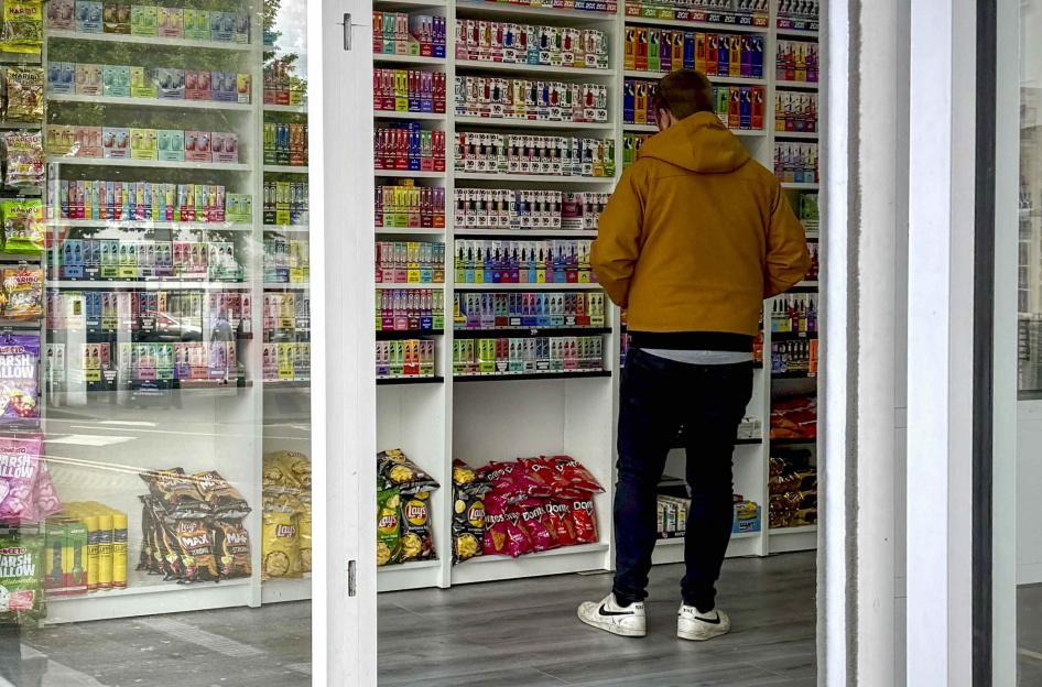 A person facing shelves filled with brightly colored vapes and other tobacco products, with snacks like chips visible at the bottom.