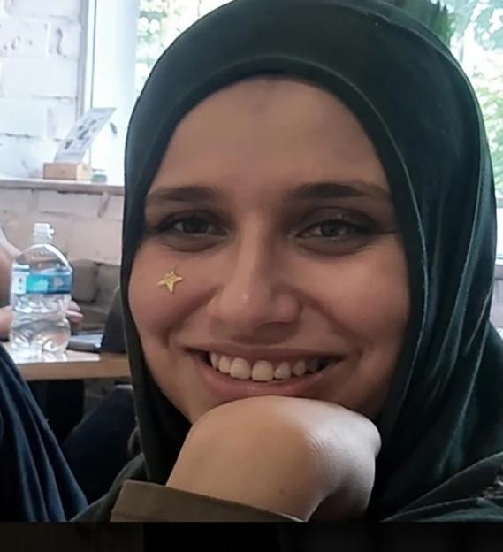 Close-up of Zeina Alian, wearing a dark green hijab and a gold star sticker on her cheek, smiling with her chin resting on her hand.