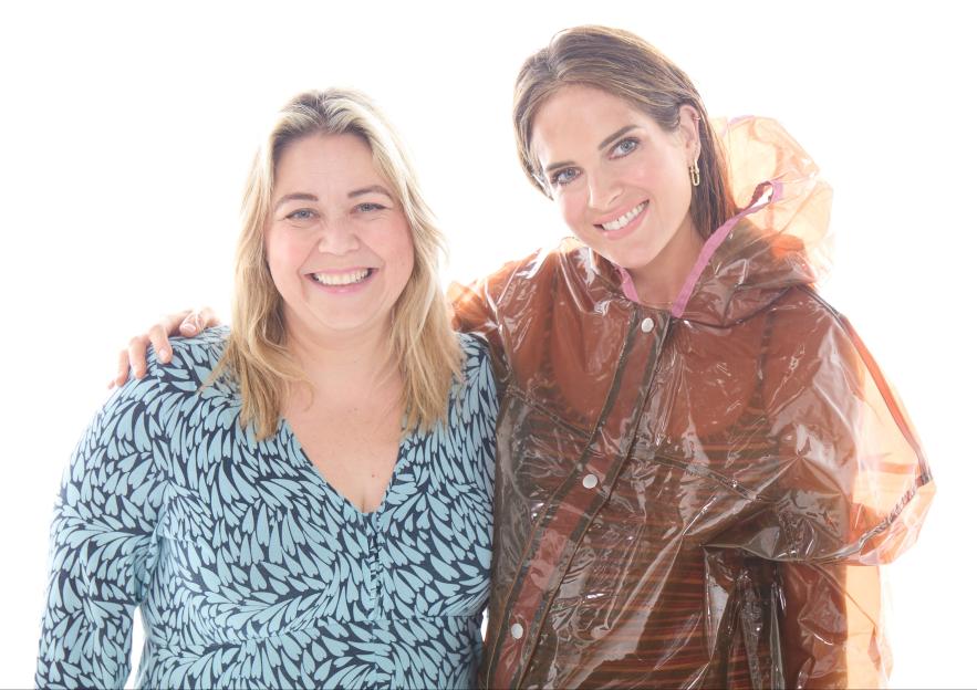 Two women smile at the camera, one with blonde hair and a blue patterned top, the other with brown hair in a clear raincoat.