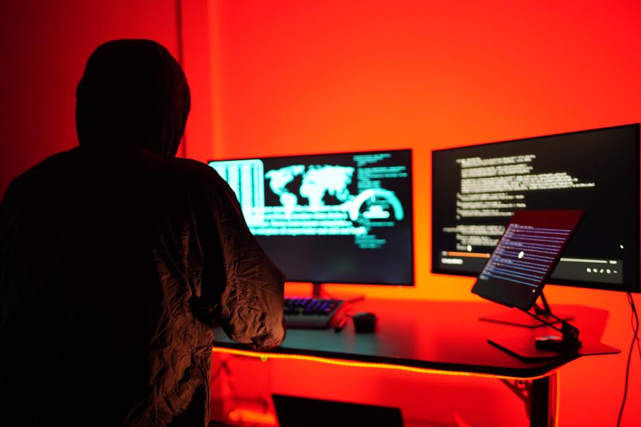 A person in a hoodie works in a dark room with a red light, illuminated by three computer screens displaying code and a world map.