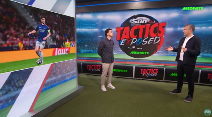 Two men discussing soccer tactics on a TV show, with one man gesturing and the other standing beside a large screen displaying a soccer player on the field.