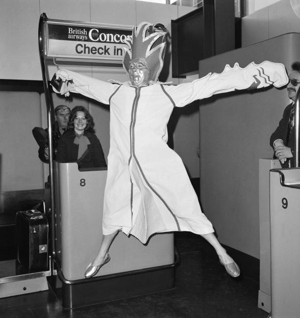 Bob Ingham, dressed in a festive jumpsuit and hat, jumps with arms outstretched at a British Airways Concorde check-in counter.