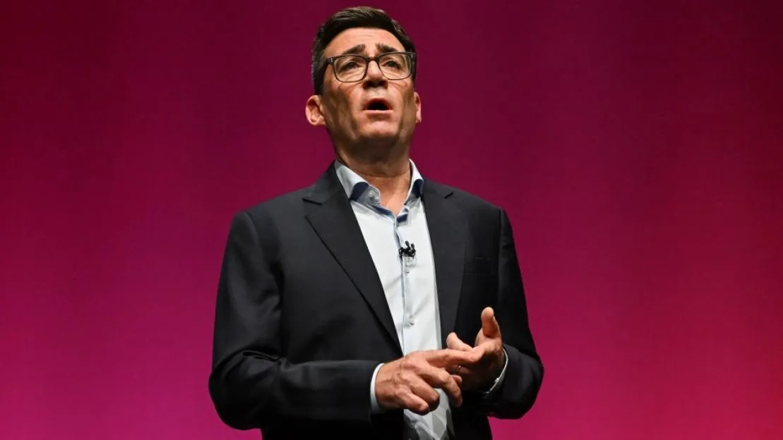 British Prime Minister Andy Burnham gives a speech on stage against a red backdrop.