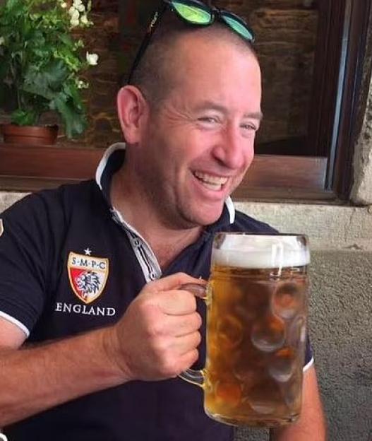 A smiling man in a dark blue polo shirt holding a large mug of beer.