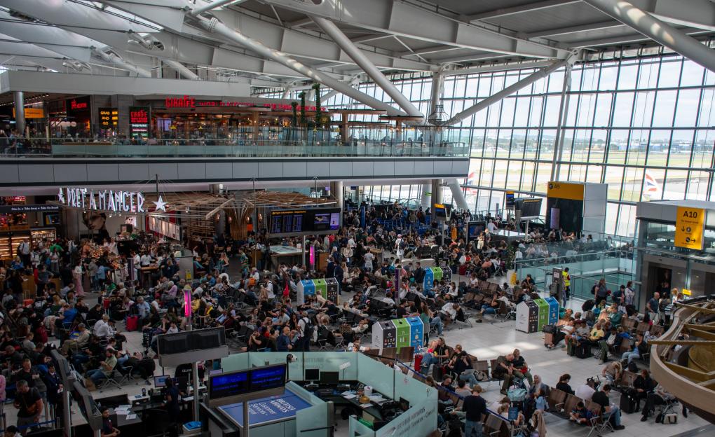 An overhead shot of a very busy Heathrow Airport, showing the large, modern interior of the airport with many people sitting and walking, and an airplane visible through the windows.