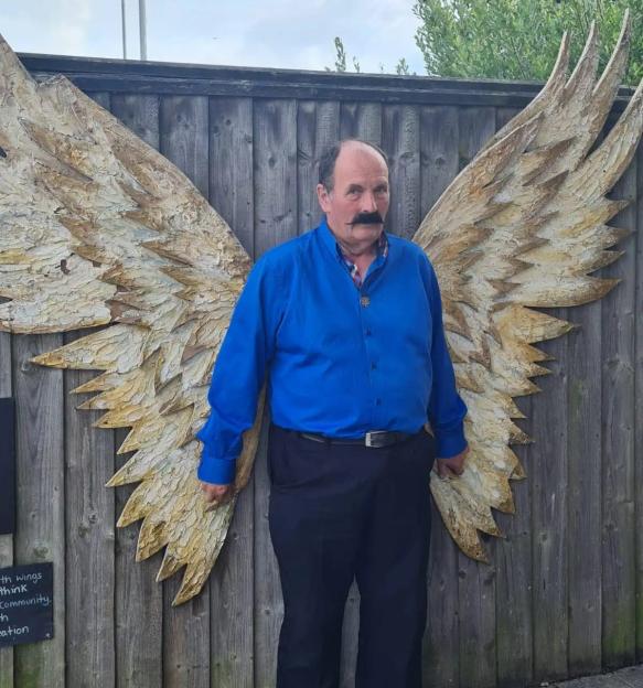 John Starkey posing in front of a wooden fence with decorative, large, golden angel wings attached.