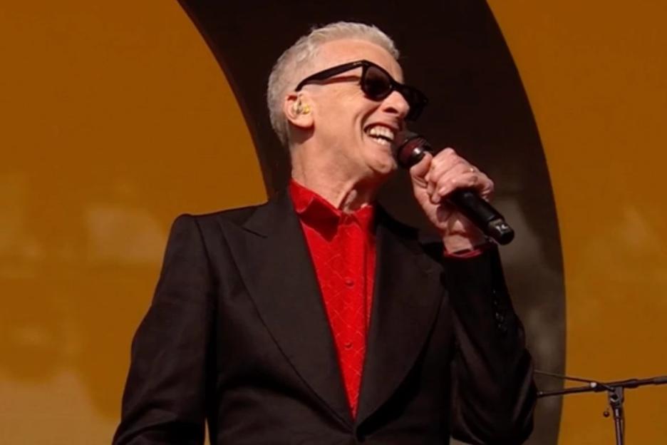 Peter Capaldi, wearing a red shirt and black suit, sings into a microphone while on stage.