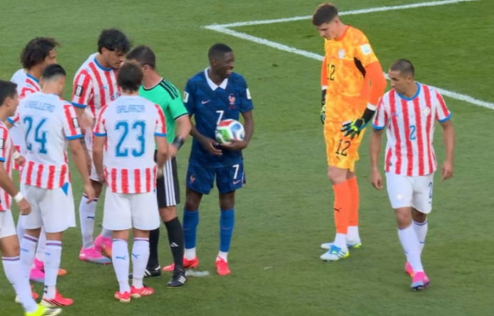Ousmane Dembele laughing while holding a football, surrounded by players in red and white striped jerseys, and an orange-clad goalkeeper.