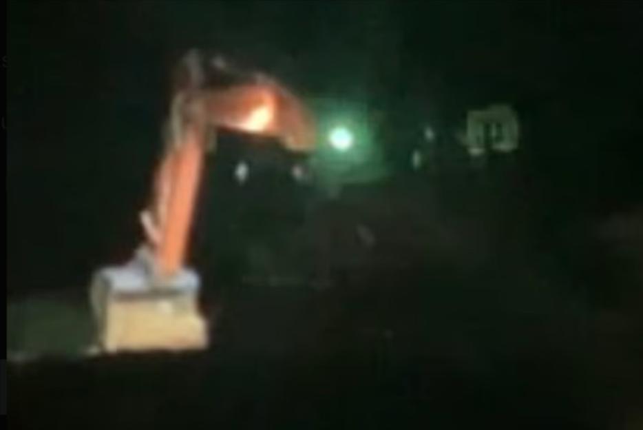 Excavator at night with a bright light in the background.