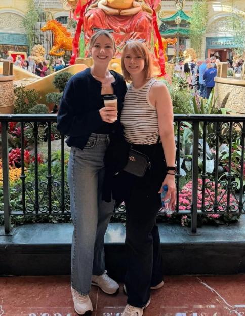 Two women smiling in front of a large golden statue of a figure holding a pot, surrounded by lush floral displays.