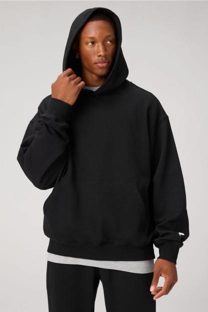 A man wearing a black hoodie with a kangaroo pocket, grey t-shirt, and black pants.