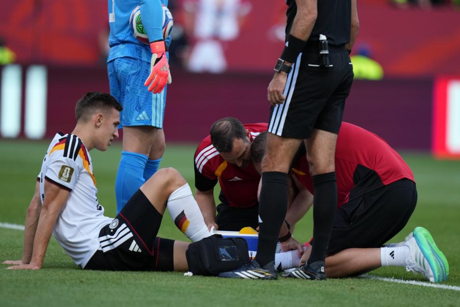 A German soccer player sitting on the field receiving medical attention from a team official, while a referee and goalkeeper stand nearby.