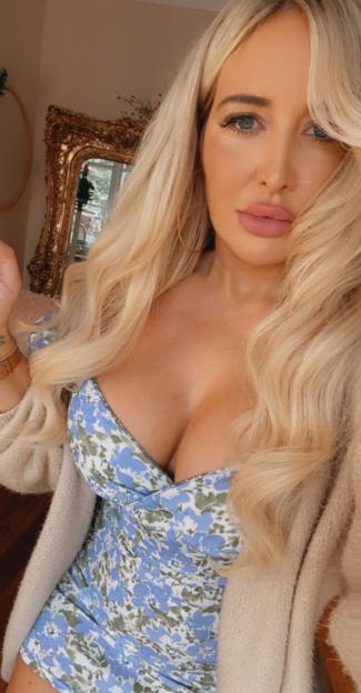 A woman with blonde wavy hair and pink lips wearing a blue floral dress and light-colored cardigan.