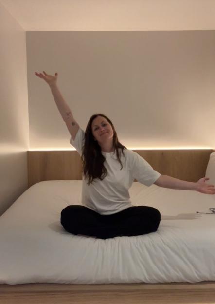 A woman sitting cross-legged on a bed, stretching with one arm raised.