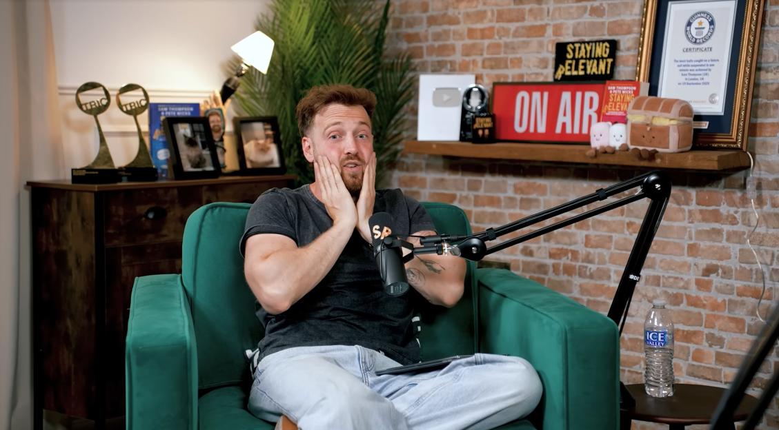 A man with short brown hair and a beard, wearing a gray t-shirt and light blue jeans, sits in a green armchair with his hands on his cheeks, appearing surprised.