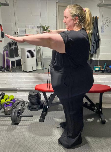 Woman in black workout clothes performing arm stretches in a gym.