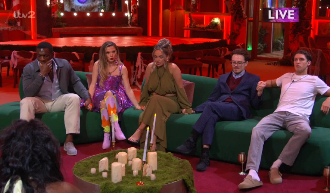 Five housemates sitting together on a green sofa, holding hands in a red-lit room.