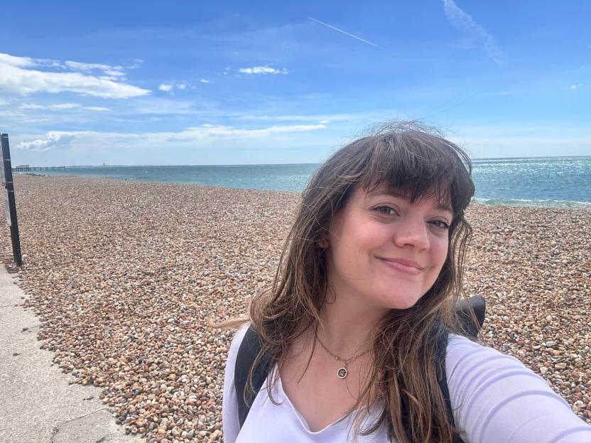 A smiling young woman in Bognor Regis, standing on a pebble beach with the sea and sky in the background.