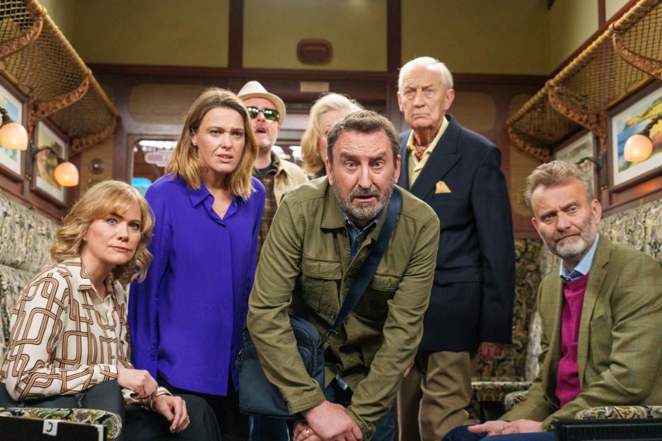 Seven people, Lee, Lucy, Martin, Wendy, Anna, Geoffrey, and Toby, look forward with concerned expressions on a train.