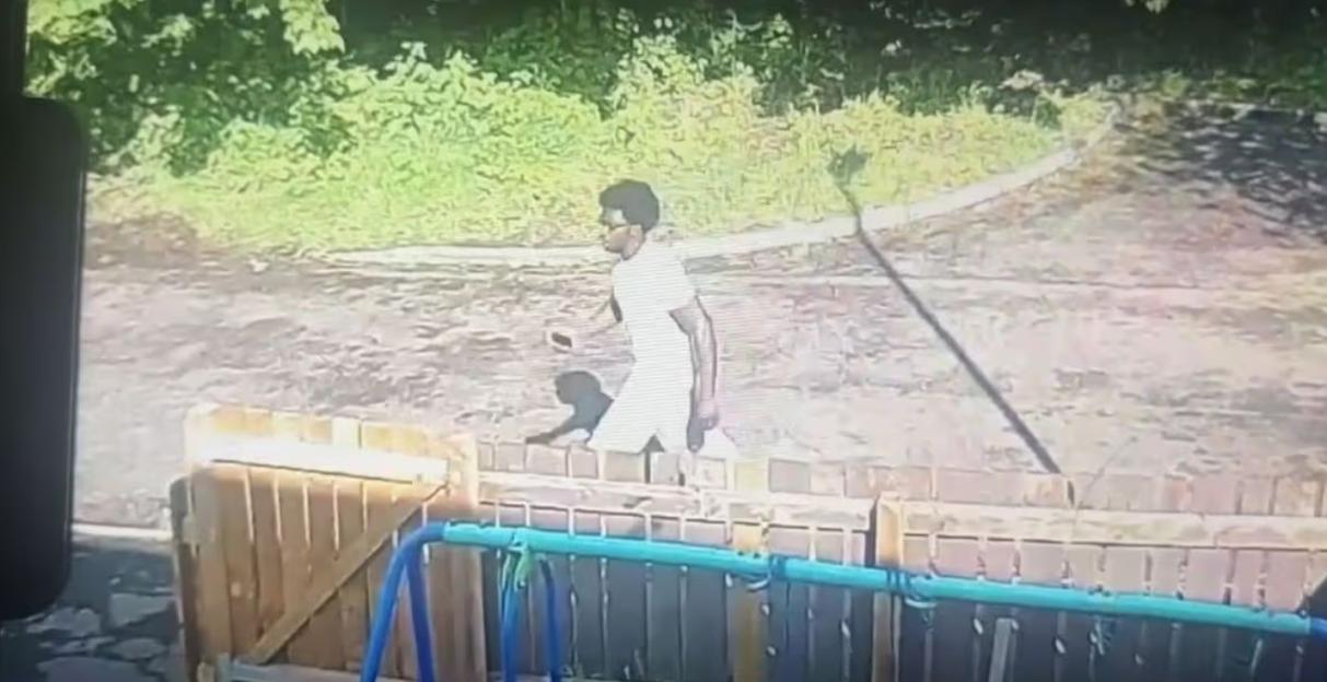 Security footage still of Gift Oladele walking past a wooden fence on a sunny day.