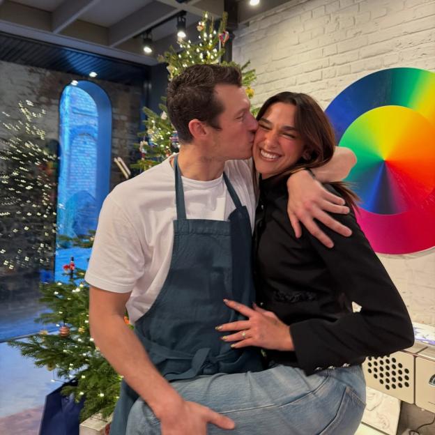 Dua Lipa sits on Callum Turner's lap as he kisses her cheek, with Christmas trees and a rainbow disc in the background.