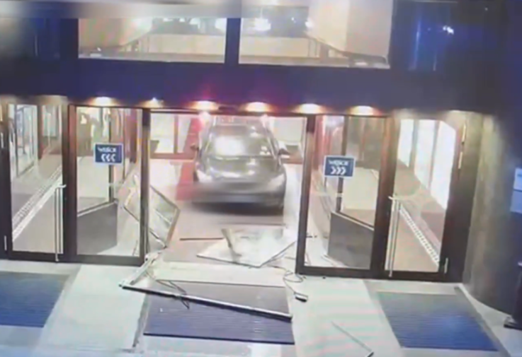 A gray car drives through the glass entrance doors of a building, shattering the glass.
