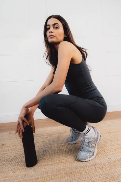 A woman in black activewear squats on a woven rug, holding a black water bottle.